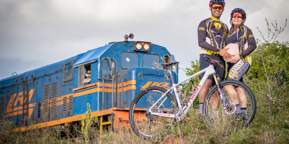 A fotografia captura 4 pessoas. É um momento de lazer ou esporte, com um homem e uma mulher vestidos com uniformes de ciclismo, ambos sorrindo e posando para a foto. Eles estão vestidos de forma semelhante, com roupas que apresentam um padrão de cores preto, amarelo e detalhes em branco, provavelmente indicando uma equipe ou clube de ciclismo. O homem usa óculos de sol com armação vermelha e um capacete de ciclismo que combina com as cores do uniforme, enquanto a mulher tem óculos de sol e o mesmo estilo de capacete. Eles estão segurando uma bicicleta de montanha com o quadro branco e detalhes em vermelho e preto, a qual está inclinada entre eles. Ao fundo, vemos uma locomotiva de trem azul com listras laranjas e brancas, com um maquinista visível na janela. A locomotiva parece ser antiga ou de um modelo mais clássico, e está em um ambiente com vegetação natural, em uma área rural. A luz do dia é suave, indicando que pode ser no fim da tarde ou em um dia parcialmente nublado. A cena transmite um sentimento de alegria e companheirismo, com um toque de aventura graças à presença do trem. E a mulher está grávida.