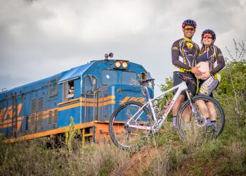 A fotografia captura 4 pessoas. É um momento de lazer ou esporte, com um homem e uma mulher vestidos com uniformes de ciclismo, ambos sorrindo e posando para a foto. Eles estão vestidos de forma semelhante, com roupas que apresentam um padrão de cores preto, amarelo e detalhes em branco, provavelmente indicando uma equipe ou clube de ciclismo. O homem usa óculos de sol com armação vermelha e um capacete de ciclismo que combina com as cores do uniforme, enquanto a mulher tem óculos de sol e o mesmo estilo de capacete. Eles estão segurando uma bicicleta de montanha com o quadro branco e detalhes em vermelho e preto, a qual está inclinada entre eles. Ao fundo, vemos uma locomotiva de trem azul com listras laranjas e brancas, com um maquinista visível na janela. A locomotiva parece ser antiga ou de um modelo mais clássico, e está em um ambiente com vegetação natural, em uma área rural. A luz do dia é suave, indicando que pode ser no fim da tarde ou em um dia parcialmente nublado. A cena transmite um sentimento de alegria e companheirismo, com um toque de aventura graças à presença do trem. E a mulher está grávida.