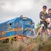 A fotografia captura 4 pessoas. É um momento de lazer ou esporte, com um homem e uma mulher vestidos com uniformes de ciclismo, ambos sorrindo e posando para a foto. Eles estão vestidos de forma semelhante, com roupas que apresentam um padrão de cores preto, amarelo e detalhes em branco, provavelmente indicando uma equipe ou clube de ciclismo. O homem usa óculos de sol com armação vermelha e um capacete de ciclismo que combina com as cores do uniforme, enquanto a mulher tem óculos de sol e o mesmo estilo de capacete. Eles estão segurando uma bicicleta de montanha com o quadro branco e detalhes em vermelho e preto, a qual está inclinada entre eles. Ao fundo, vemos uma locomotiva de trem azul com listras laranjas e brancas, com um maquinista visível na janela. A locomotiva parece ser antiga ou de um modelo mais clássico, e está em um ambiente com vegetação natural, em uma área rural. A luz do dia é suave, indicando que pode ser no fim da tarde ou em um dia parcialmente nublado. A cena transmite um sentimento de alegria e companheirismo, com um toque de aventura graças à presença do trem. E a mulher está grávida.